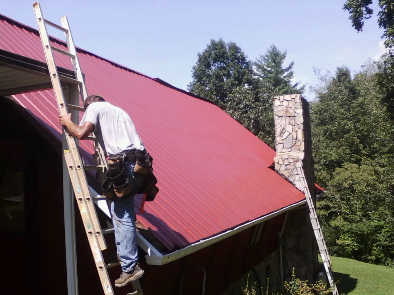 Roofer on a ladder beside a red metal roof and stone chimney — Soffit & Fascia Repair by Roofing Brothers in Dearborn